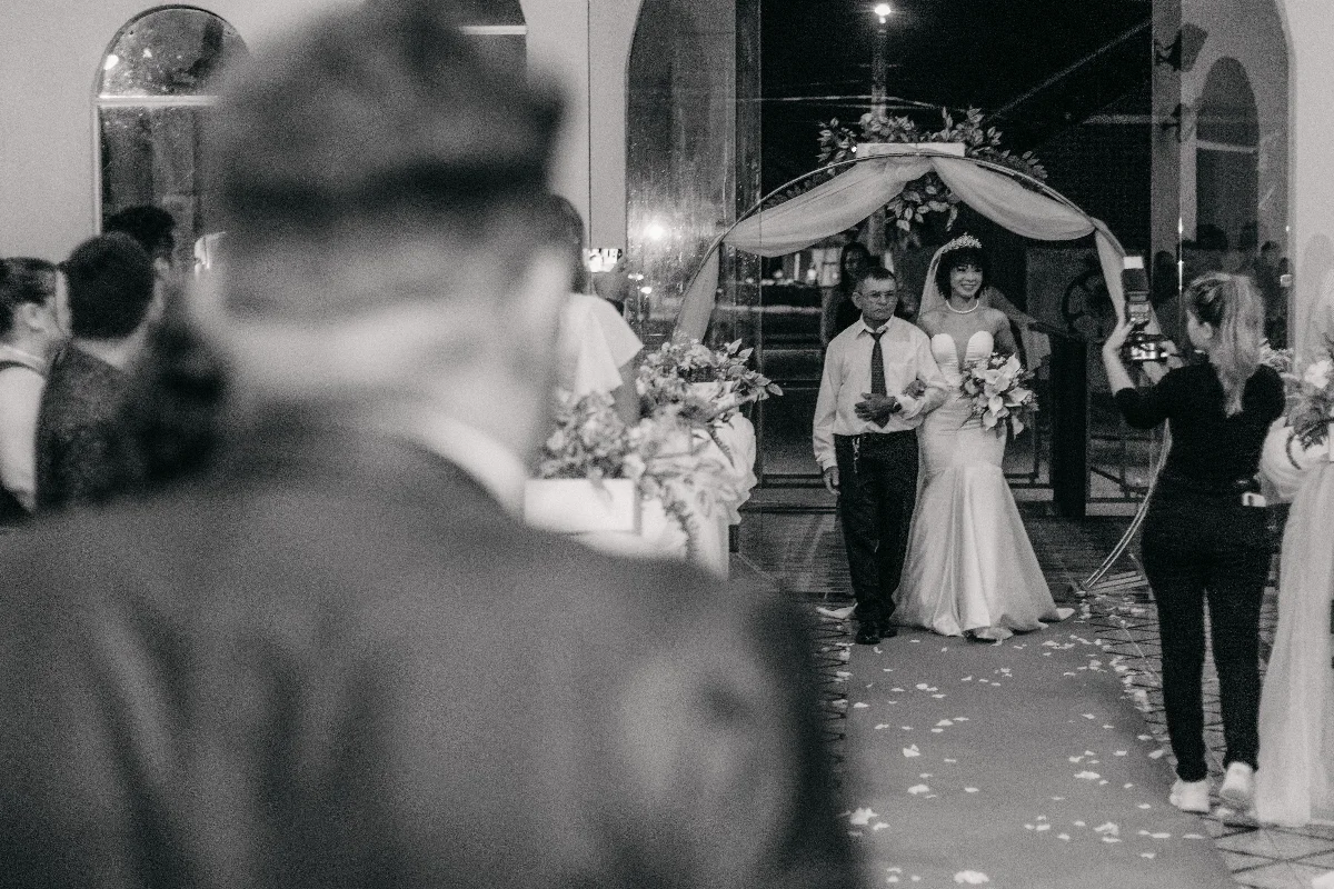 Father walks smiling bride down the aisle at indoor wedding ceremony. Captured in black and white.
