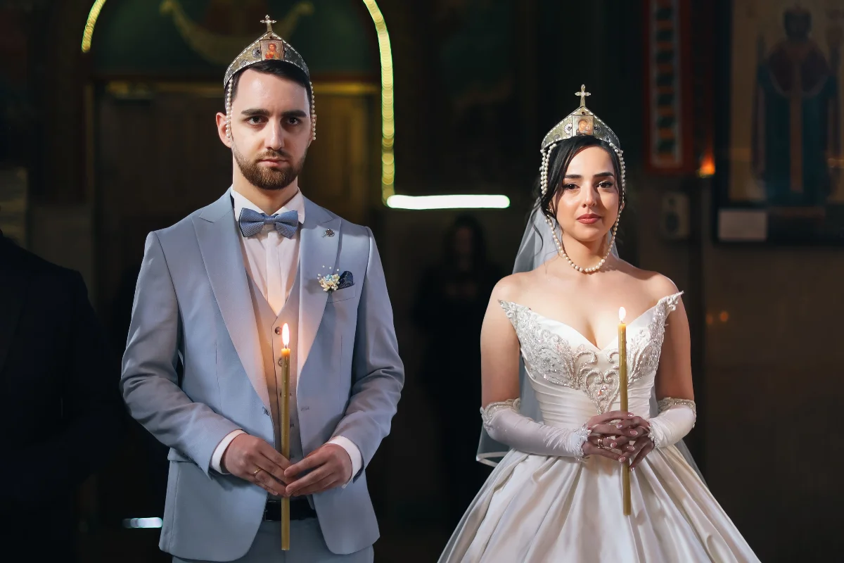 Bride and groom in traditional attire holding candles during a church wedding ceremony.
