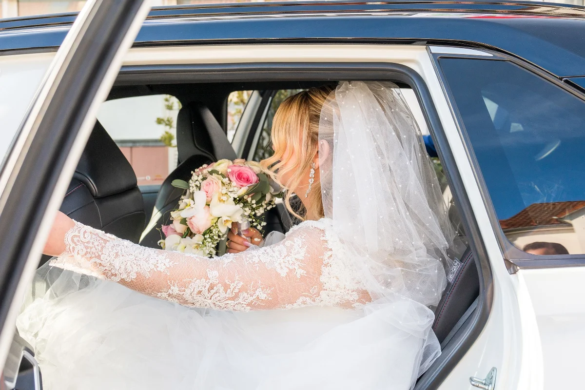 Elegant bride in lace wedding dress and veil holding a bouquet inside a car.