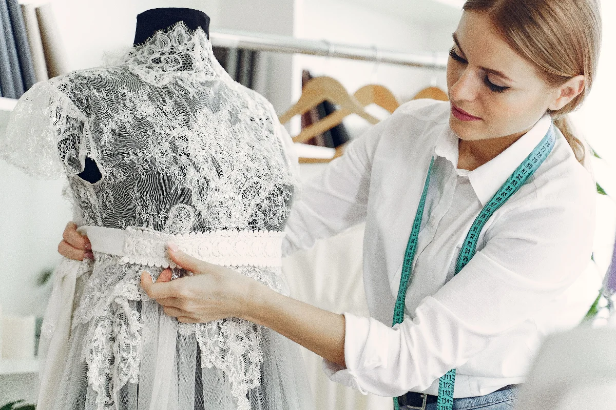 Talented female tailor wearing white shirt and jeans working with mannequin in modern atelier or wedding boutique while attaching belt on lace dress