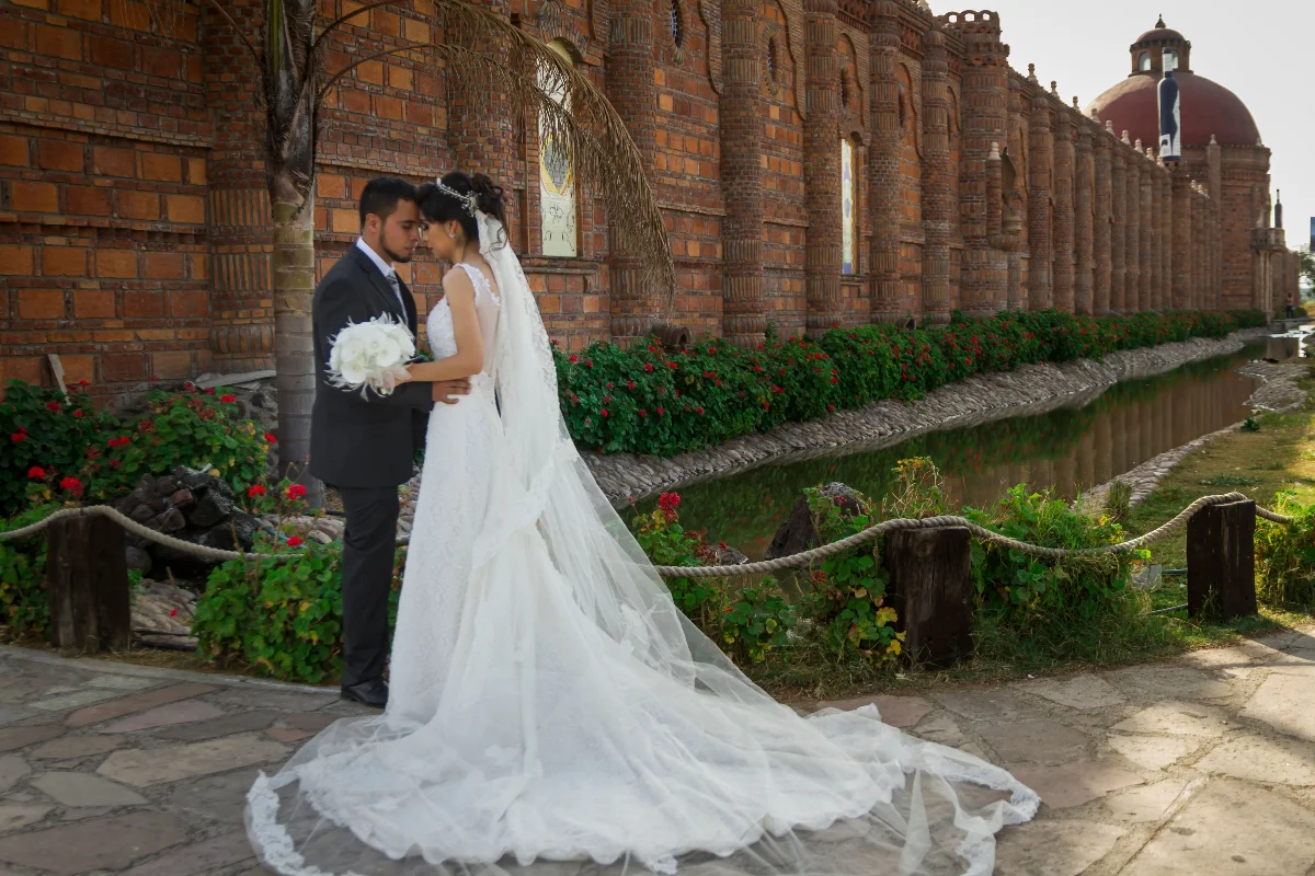 Bride and groom share a tender moment by a historic brick venue, surrounded by vibrant greenery.