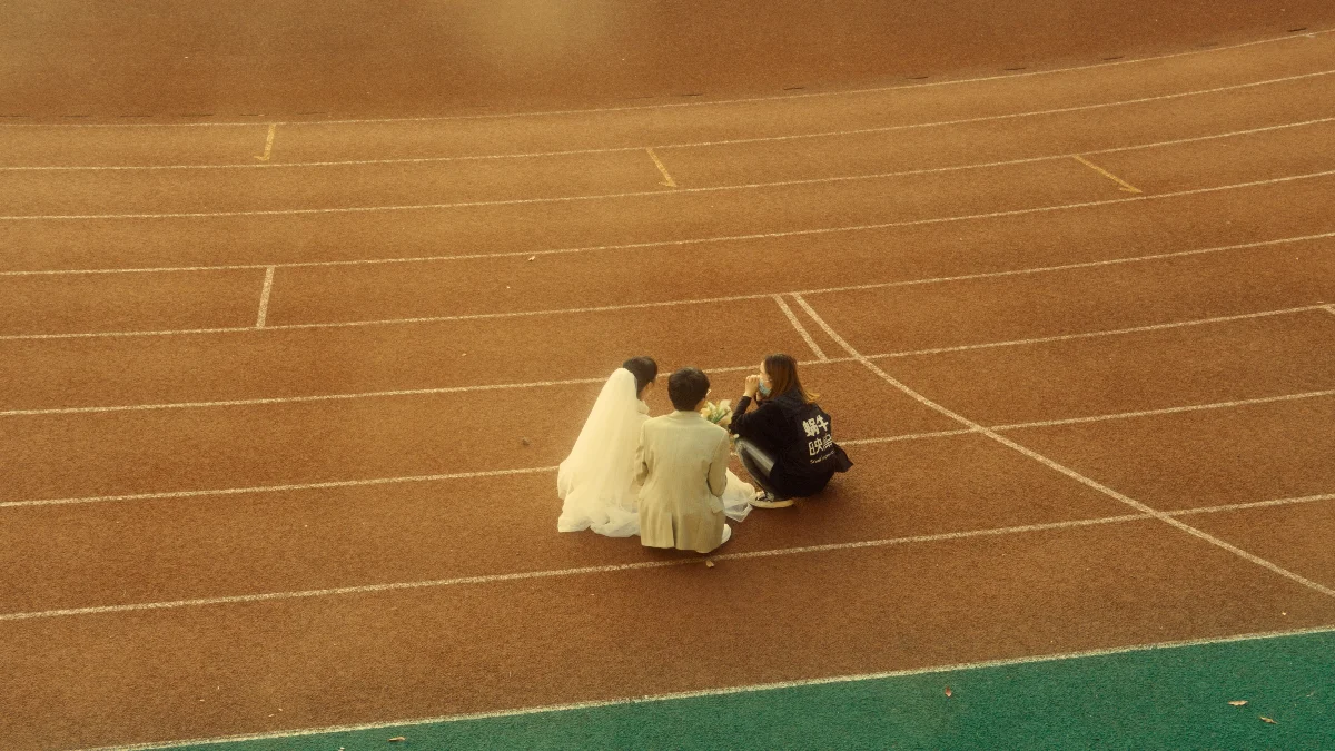 A bride and groom in wedding attire sitting together on a sports running track.
