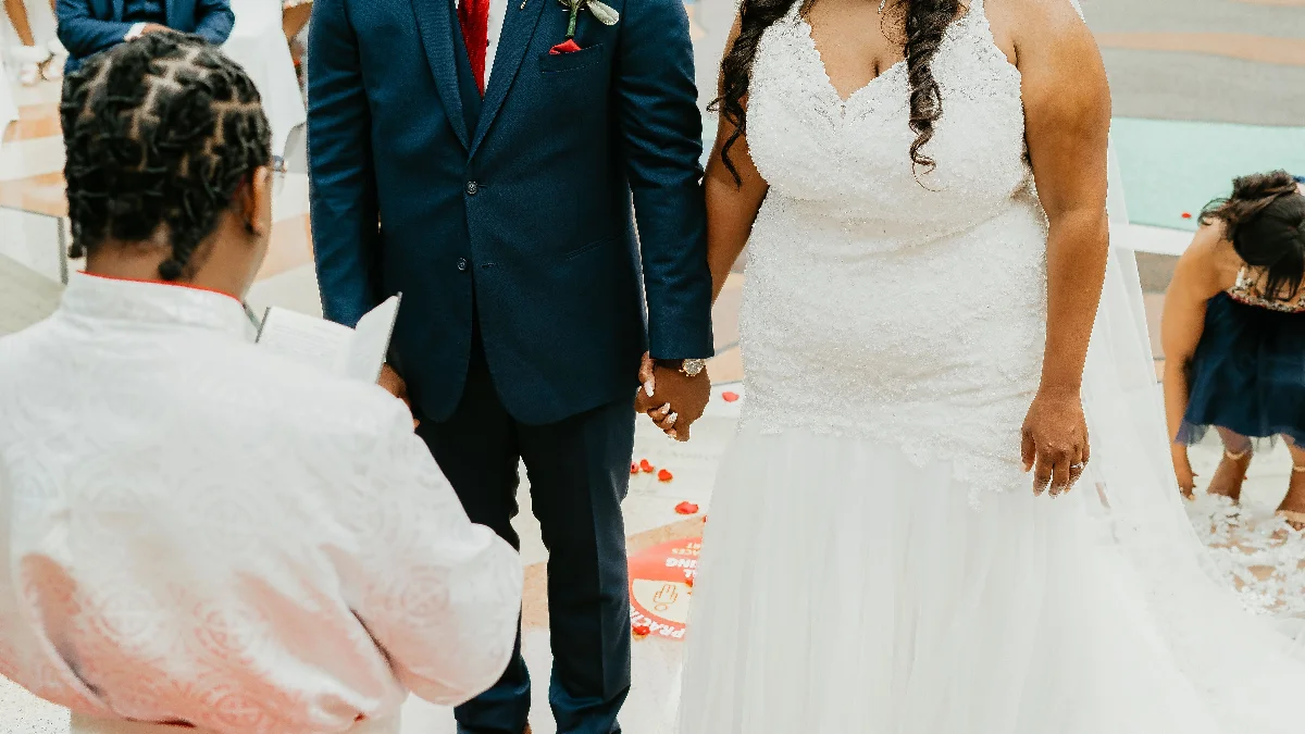 Crop anonymous bride and groom in elegant wedding outfits holding hands while standing on street with guests during festive ceremony