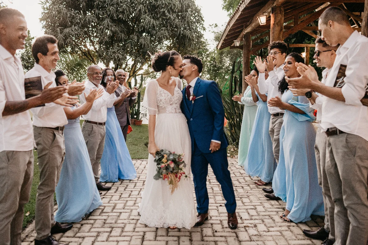 Young bride with flower bouquet kissing black husband between cheerful clapping guests on tiled walkway during festive event