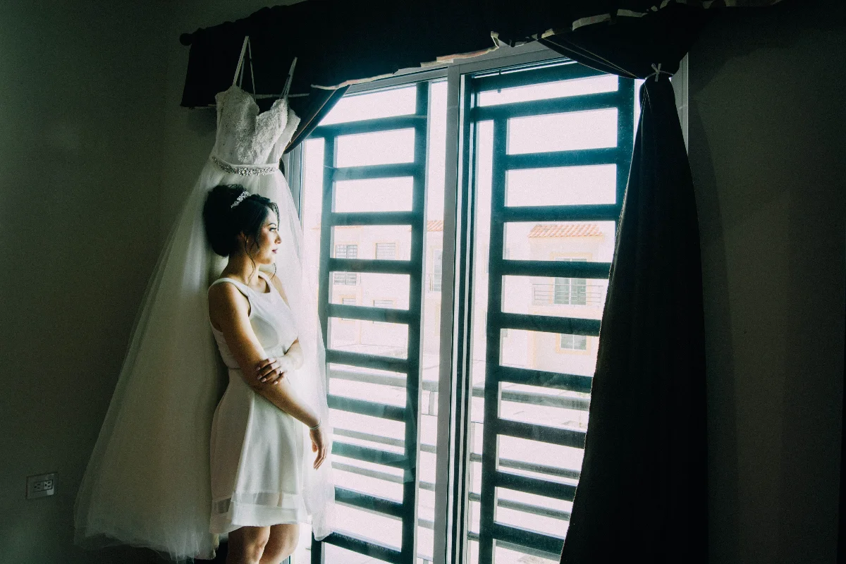 Bride stands by window in white dress, reflecting on her special day. Captures intimate wedding moment.