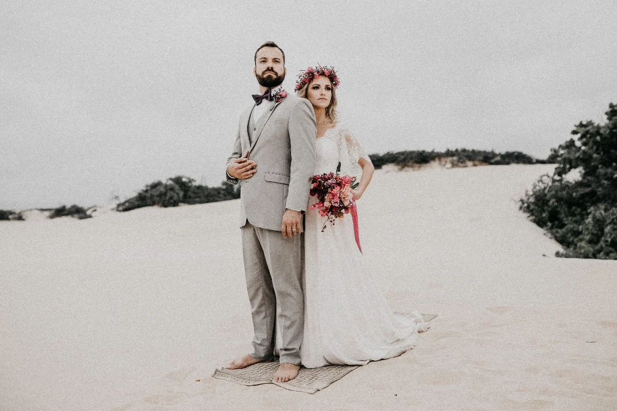 Bride and groom in a romantic setting on sand dunes wearing elegant attire.