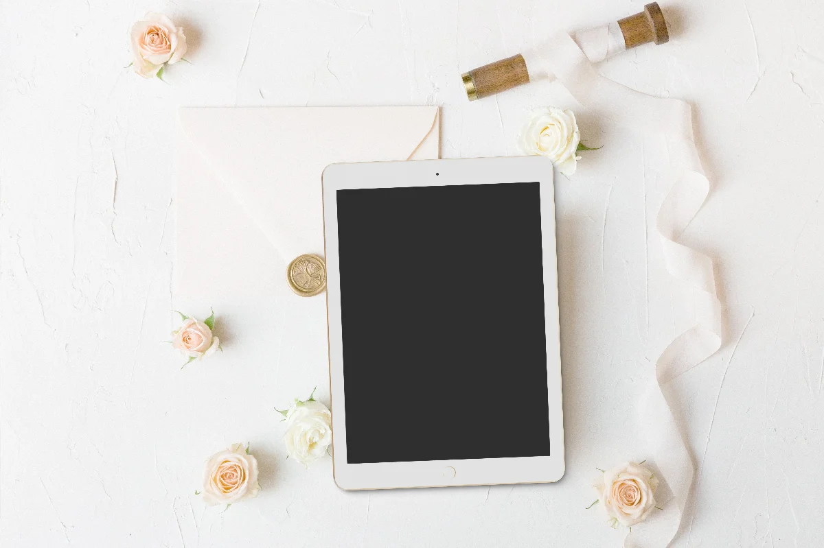 Stylish flat lay featuring a digital tablet surrounded by roses and an envelope on a white background.