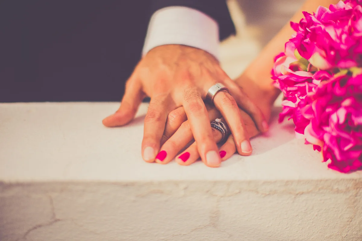 Close-up of a married couple's hands with wedding rings and pink flowers, symbolizing love and commitment.